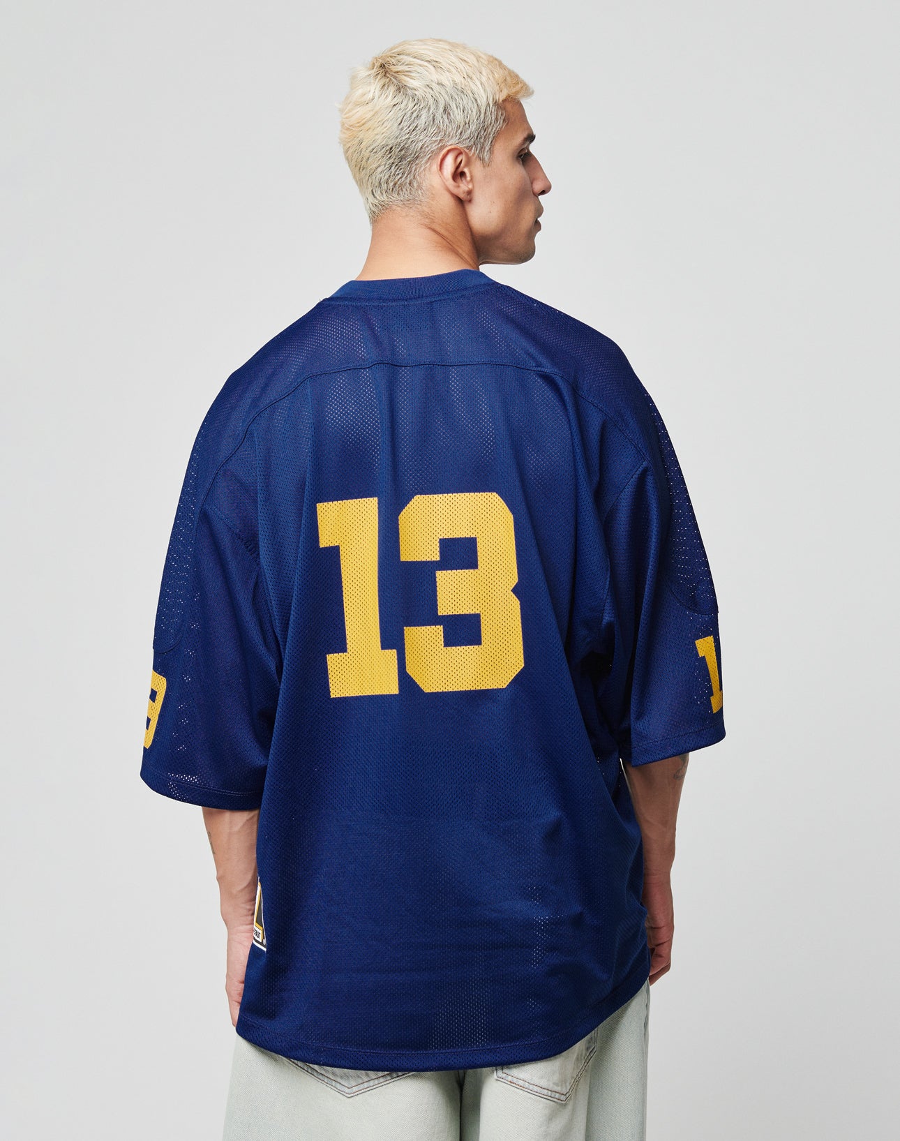 A person with short, bleached blonde hair wears the oversize LFDY Mesh Jersey by LIVE FAST DIE YOUNG. The loose-fit blue polyester jersey features a V-neck and yellow number 13 on the back and sleeves. The person faces away from a plain background.