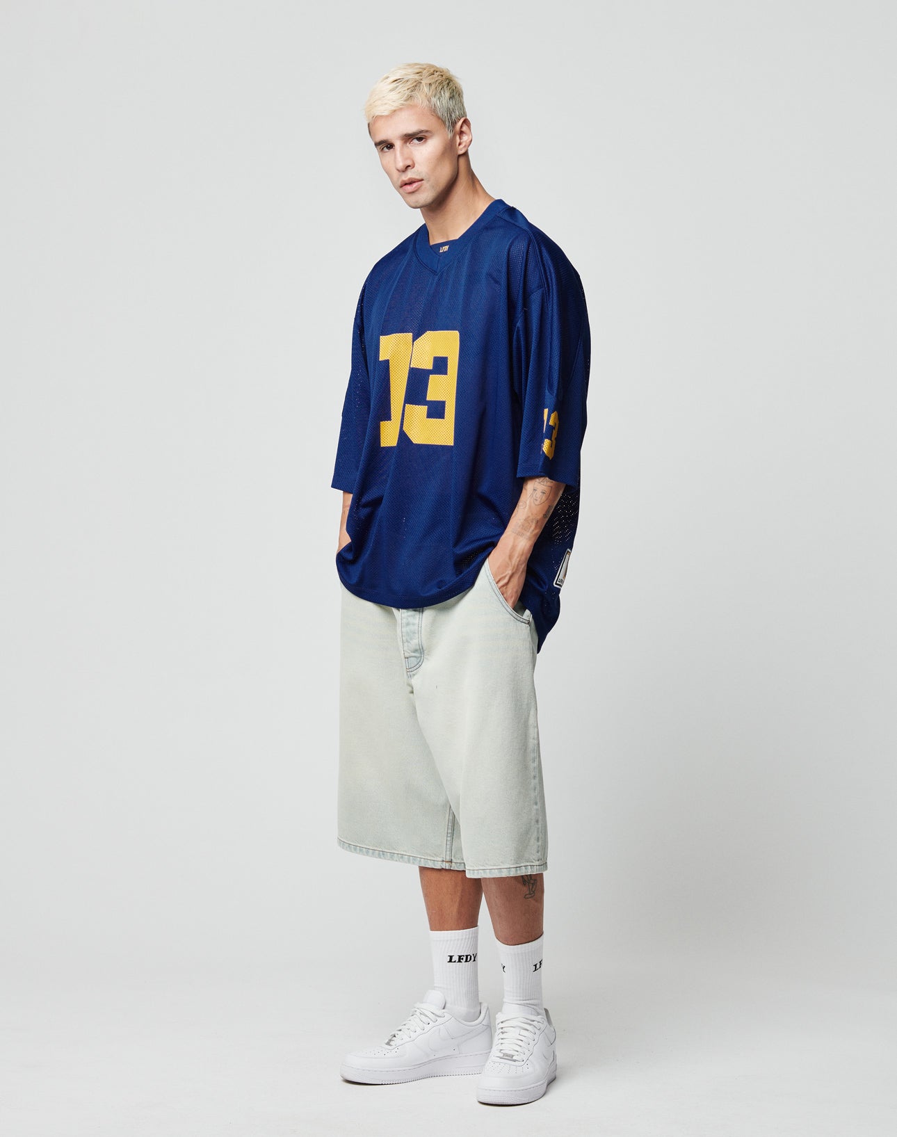 A man with short blond hair wears an oversize fit blue LFDY Mesh Jersey by LIVE FAST DIE YOUNG, light baggy shorts, white socks, and white sneakers. He stands with hands in pockets against a plain white background.