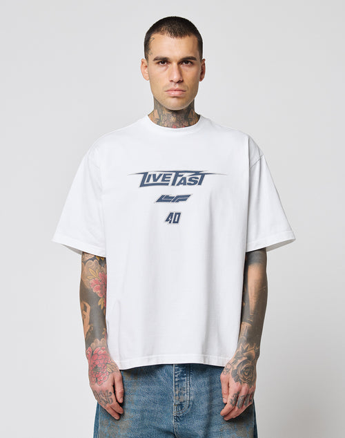 A tattooed man wears the oversize fit LFDY Triple Racing Tee by LIVE FAST DIE YOUNG, a white 100% Baumwolle shirt with LIVE FAST and 40 in blue, paired with blue jeans. He stands against a plain light background, facing the camera.