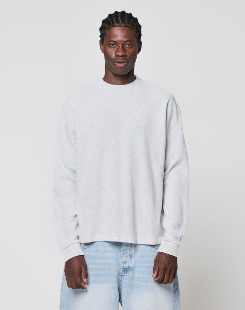 A man with braided hair wears the LIVE FAST DIE YOUNG Blank Waffle Longsleeve in light gray and light blue jeans, standing against a plain white background and looking straight ahead with a neutral expression.