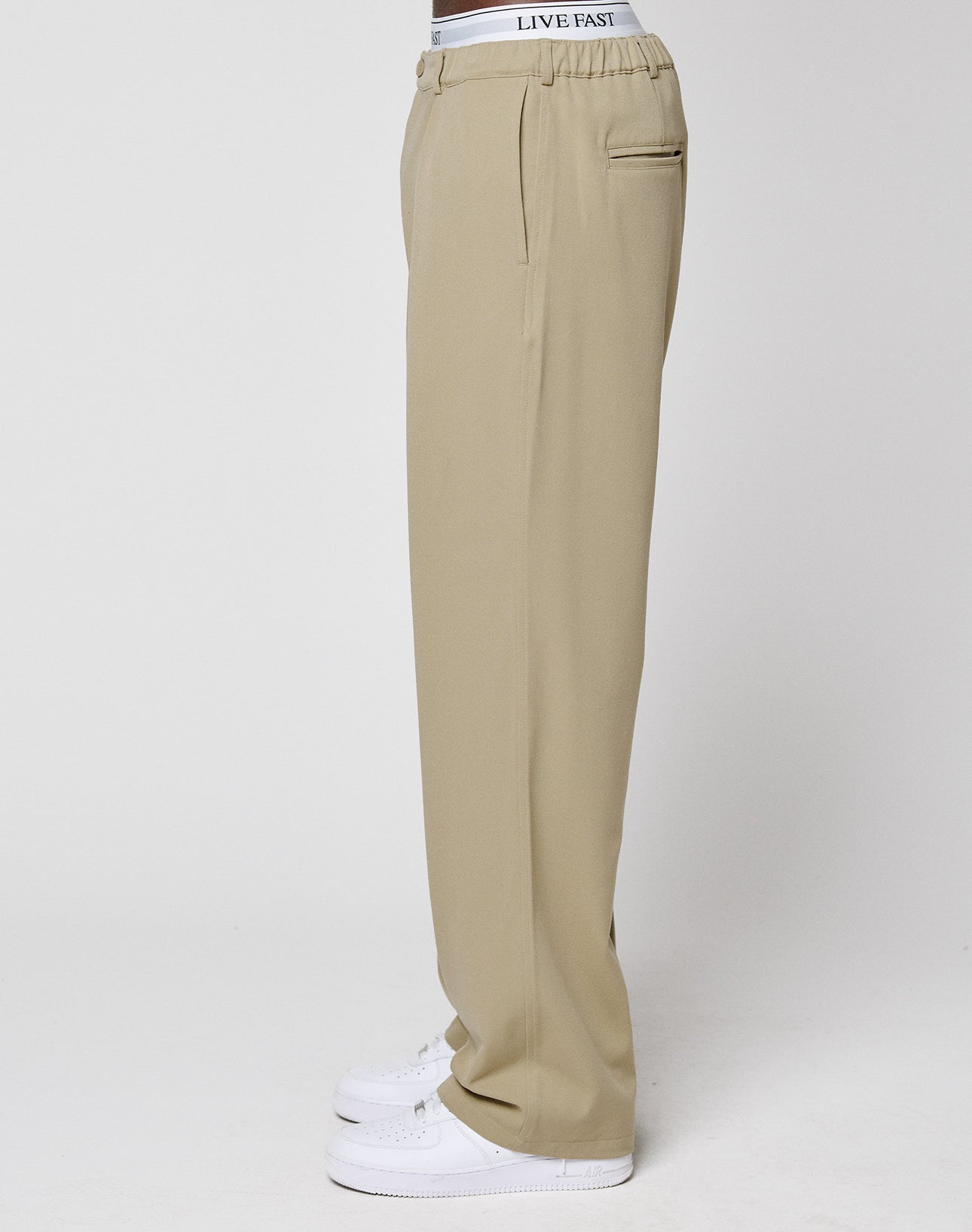 A person stands sideways against a plain white background, wearing LIVE FAST DIE YOUNG Wide Casual Pants in beige with wide legs, a visible white waistband featuring "LIVE FAST" text, and white sneakers.