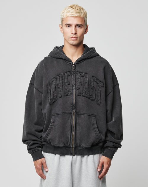 A young man with short blond hair wears the LIVE FAST DIE YOUNG Ripped College Hooded Zip, a loose black hoodie with textured LIVE FAST on the front, made from 100% Baumwolle, and light gray sweatpants against a plain gray background.
