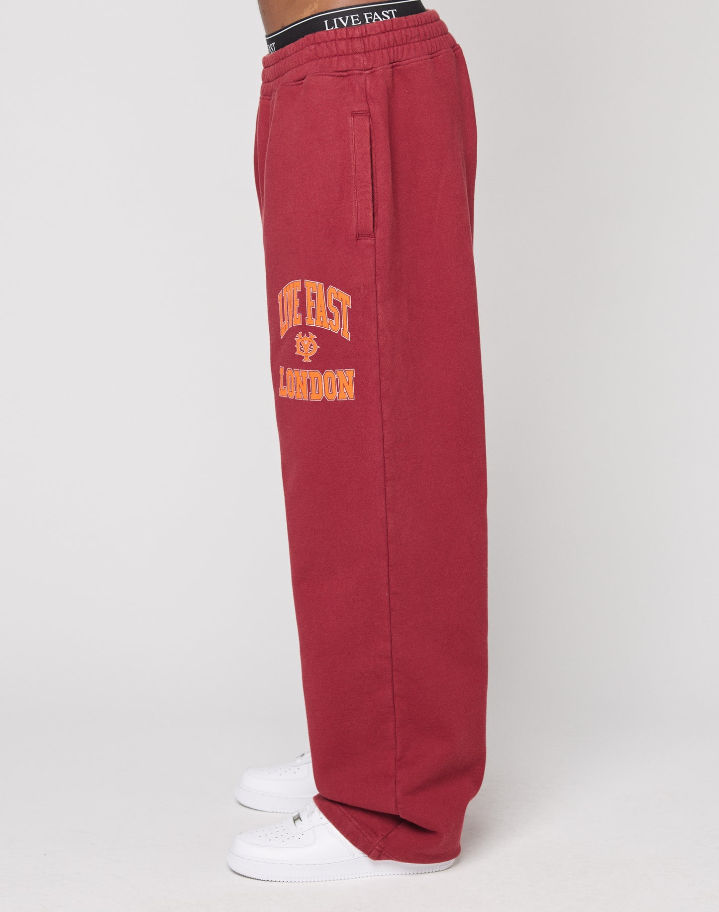 A person stands sideways wearing LIVE FAST DIE YOUNG’s Live Fast London Wide Jogger in red cotton with yellow “LIVE FAST LONDON” text and graphic on the left thigh, white sneakers, and an elastic waistband that reads LIVE FAST. Plain white background.