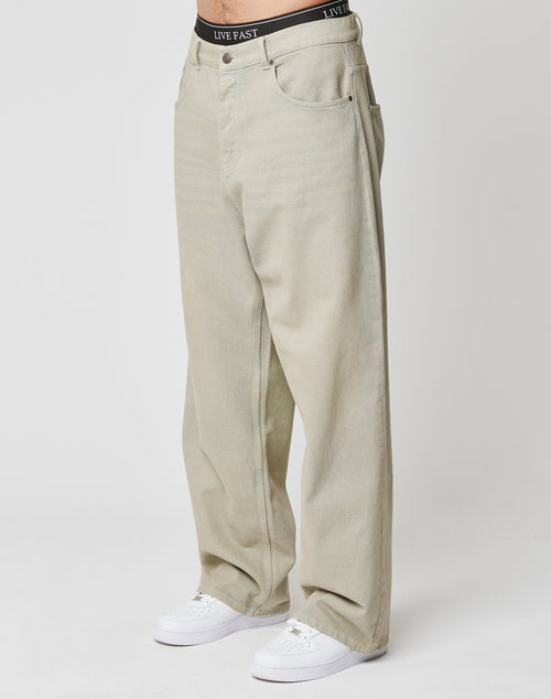 A person wears LFDY Baggy Denim by LIVE FAST DIE YOUNG—loose-fitting, light beige jeans with a visible black waistband featuring LIVE FAST text. Styled with white sneakers, made from hochwertige Baumwolle, against a plain background.