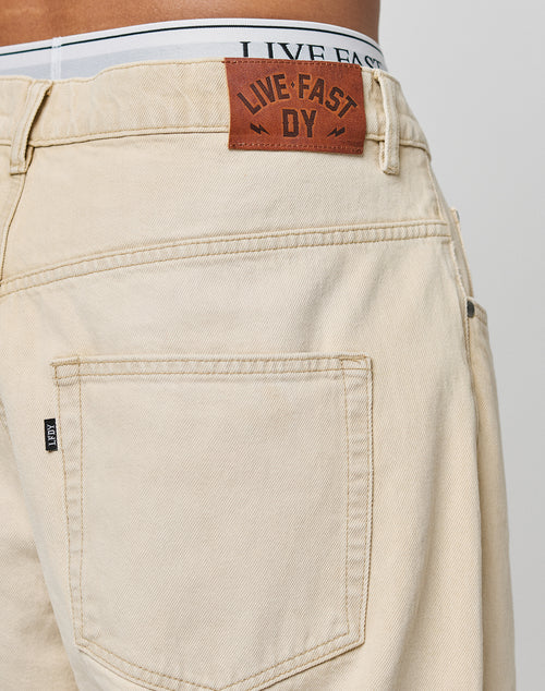 A close-up of the back of the LFDY Baggy Denim by LIVE FAST DIE YOUNG shows beige denim in a baggy fit, a brown leather patch with LIVE FAST D Y, and part of a white waistband displaying LIVE FAST.