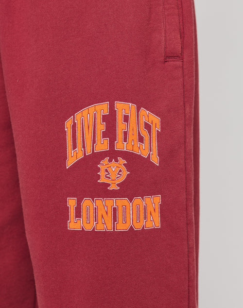 Close-up of the Live Fast London Wide Jogger by LIVE FAST DIE YOUNG: maroon wide-fit sweatpants with bold yellow and orange LIVE FAST LONDON text and emblem on the left thigh. Made from soft cotton with an elastic waist for comfort.
