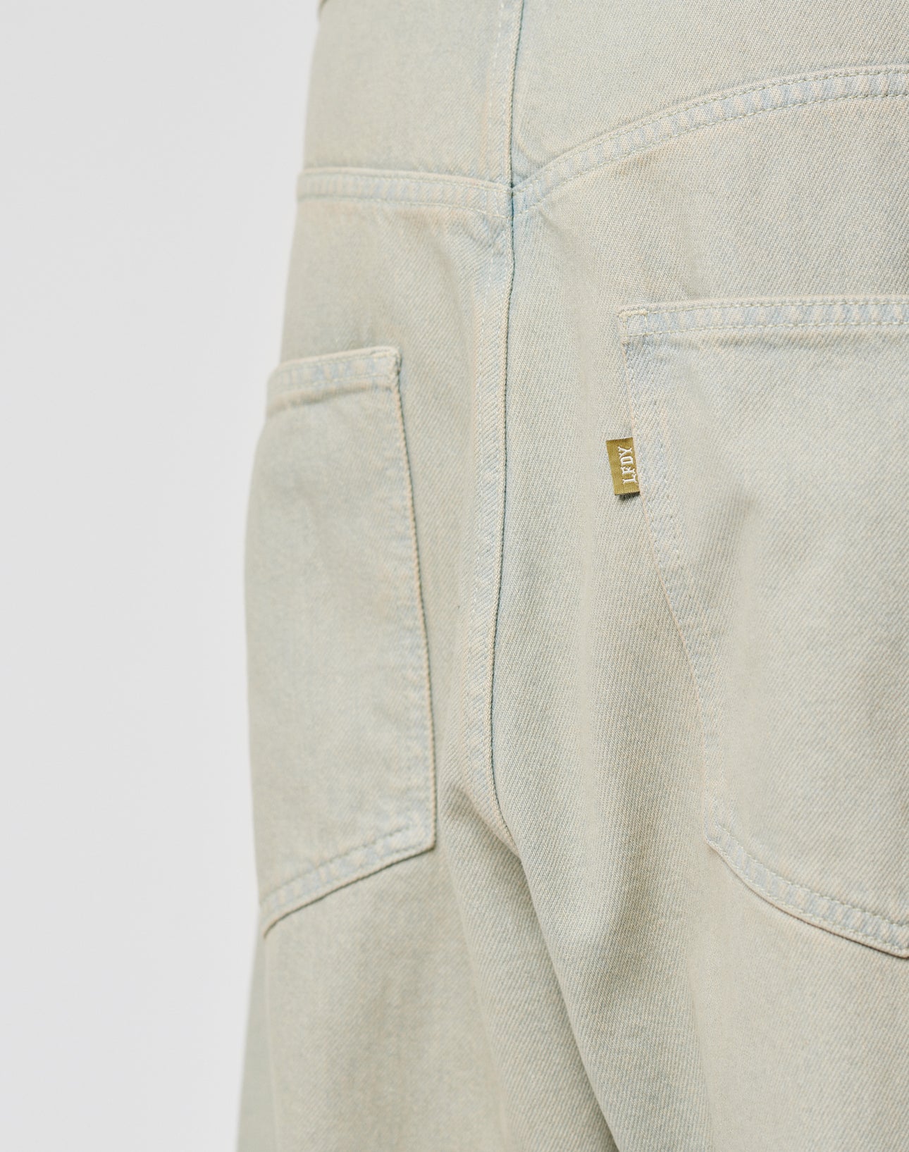 Close-up of the LIVE FAST DIE YOUNG LFDY Baggy Denim shows two back pockets, a gold-tone flag-label on the right pocket, and visible seams and stitching on light-colored baggy-fit jeans.