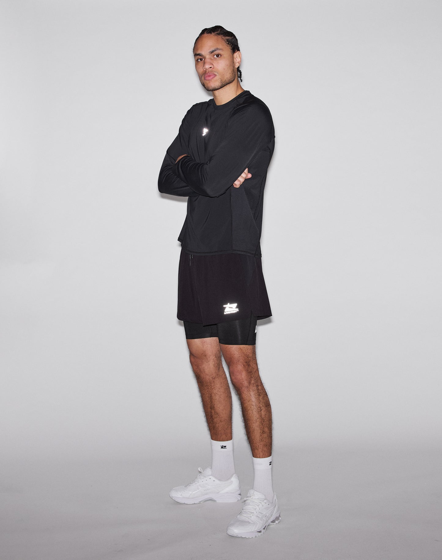 A person stands with arms crossed, wearing a black Regular Fit long-sleeve shirt, LIVE FAST DIE YOUNG Live Fast Performance Running Shorts in black with integrated tights, white socks, and white sneakers against a light-gray background.