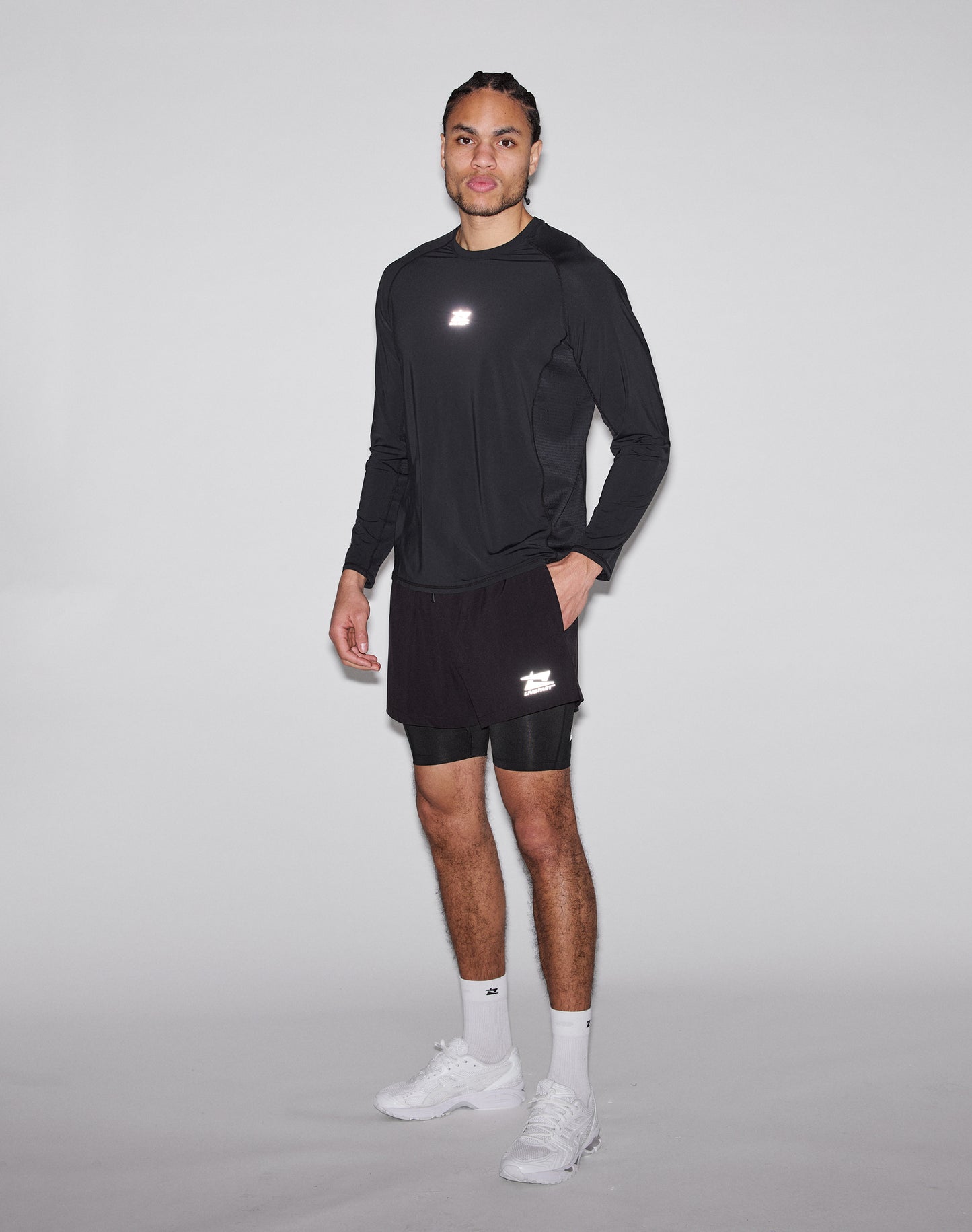 A man poses against a plain background, wearing the LIVE FAST DIE YOUNG Live Fast Performance Longsleeve in black with a reflective front print, black shorts over compression shorts, plus white socks and sneakers. He gazes neutrally at the camera.
