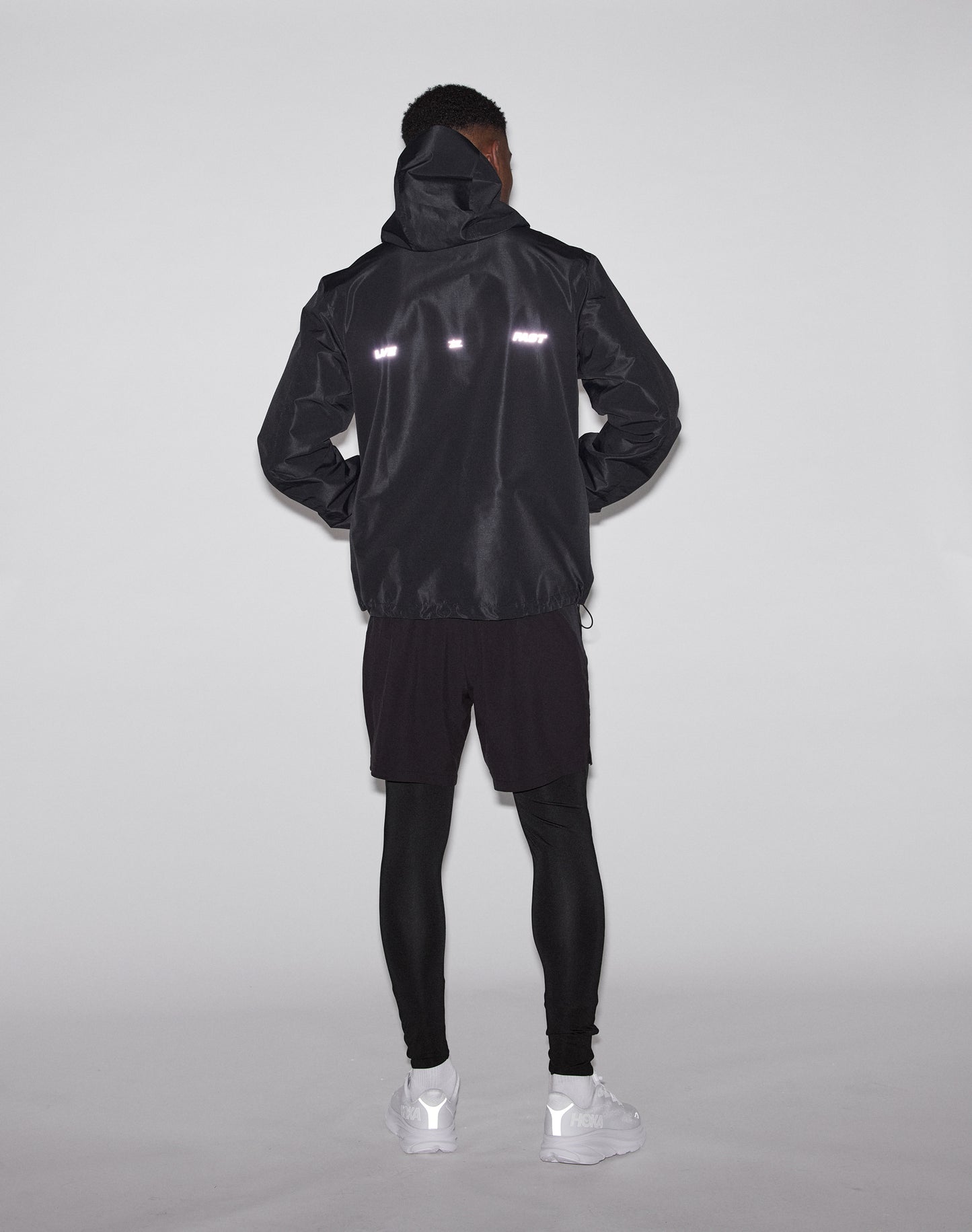 A person stands facing away, wearing the LIVE FAST DIE YOUNG Live Fast Performance Rain Jacket with waterproof seams and reflective strips, black shorts over leggings, and white sneakers against a plain light background.