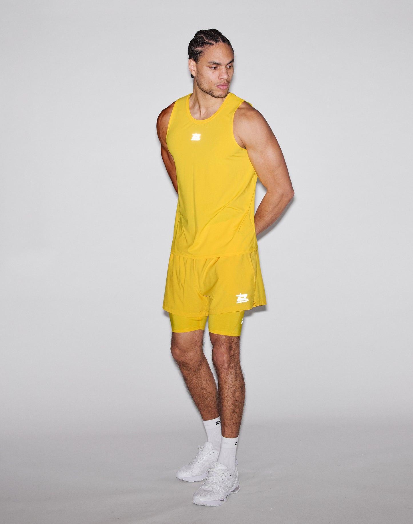 A man stands against a plain gray background wearing the LIVE FAST DIE YOUNG Live Fast Performance Tank in bright yellow and matching shorts with white sneakers, looking down to his left with his hands behind his back.