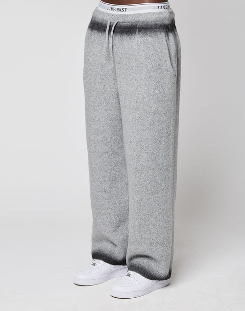 Someone wears LIVE FAST DIE YOUNG's Live Fast Boucle Pants—wide fit, light gray, straight leg with elastic waistband labeled "LIVE FAST"—with white sneakers, standing against a plain background.