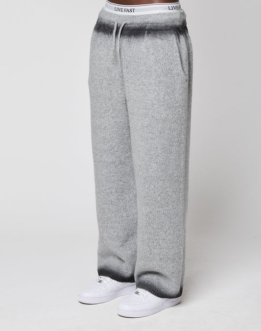 Someone wears LIVE FAST DIE YOUNG's Live Fast Boucle Pants—wide fit, light gray, straight leg with elastic waistband labeled "LIVE FAST"—with white sneakers, standing against a plain background.
