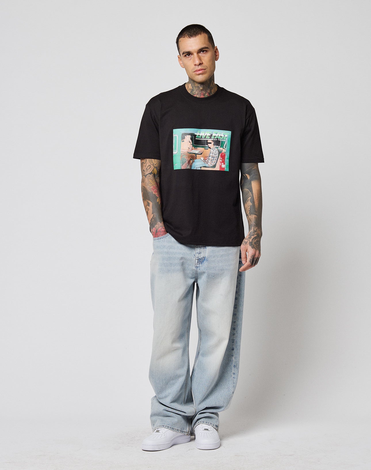 A man with tattoos wearing a black LIVE FAST DIE YOUNG LFDY Sixers Tee, light blue baggy Regular Fit jeans, and white sneakers stands against a plain white background, looking directly at the camera with a neutral expression.