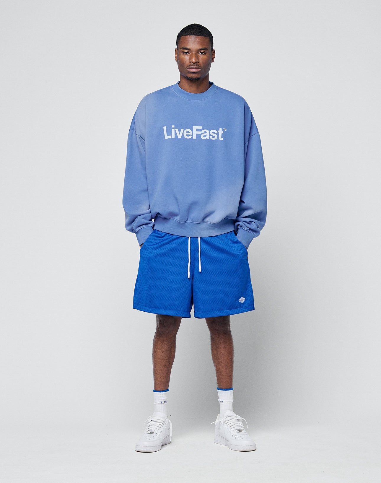 A man stands against a plain background wearing a blue LIVE FAST DIE YOUNG sweatshirt and Basic Mesh Shorts, paired with white socks and sneakers, his hands in his pockets.