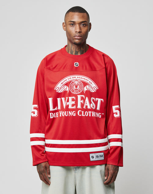 A man stands against a plain light gray background wearing an oversize fit red LFDY Hockey Jersey by LIVE FAST DIE YOUNG, featuring bold white text and the number 55 printed on both sleeves.