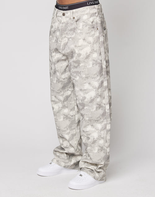 A person wears LIVE FAST DIE YOUNG’s Live Fast Canvas Pants, featuring loose-fitting light gray camouflage with a Baggy Fit and straight leg, paired with white sneakers. The waistband says LIVE FAST. The background is plain white.