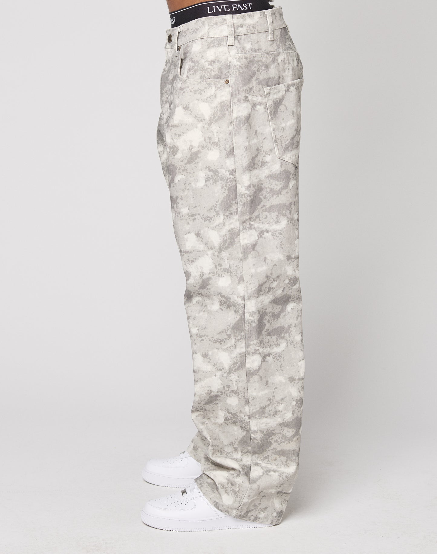 A person stands sideways in LIVE FAST DIE YOUNG’s Live Fast Canvas Pants—light gray camo with a baggy fit and wide straight leg—paired with white sneakers and a black LIVE FAST waistband, set against a plain white background.