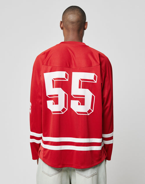 A person with a shaved head wears the LIVE FAST DIE YOUNG LFDY Hockey Jersey—an oversized red polyester top with white stripes and bold “55” print on the back—paired with light-colored jeans, facing away from the camera.