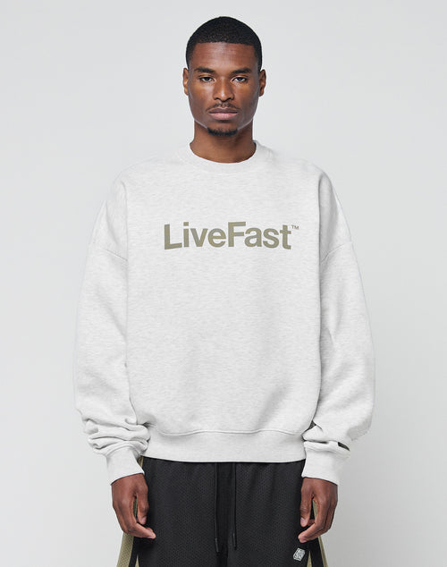 A man wearing the Basic LiveFast Sweater by LIVE FAST DIE YOUNG, a light gray oversize fit made from soft cotton with LiveFast™ on the front, stands against a plain background, looking straight at the camera with a neutral expression.