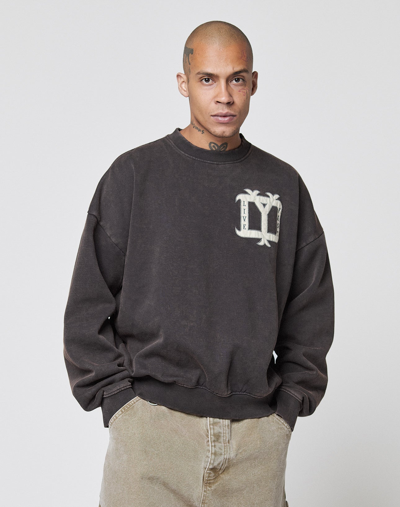 A person with a shaved head and tattoos wears a loose, dark Live Fast DY Sweater by LIVE FAST DIE YOUNG featuring a palm tree patch on the chest, paired with light pants, standing against a plain light background.