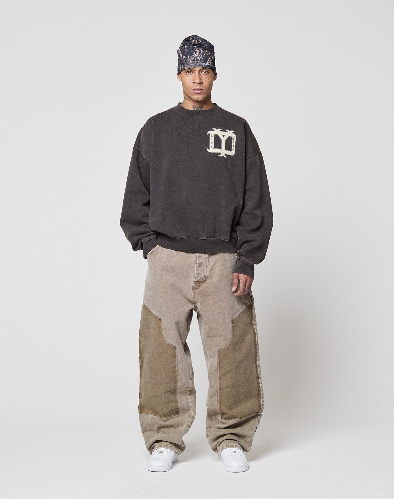 A person stands against a plain background wearing the Live Fast DY Sweater by LIVE FAST DIE YOUNG, loose light brown patchwork jeans, white sneakers, and a patterned headscarf for a subtle vintage effect.