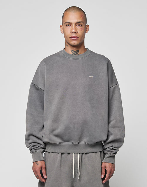 A person with a shaved head wears the LIVE FAST DIE YOUNG Basic Chest Sweater in an oversized, faded gray fit, paired with matching sweatpants and a subtle front print, standing facing forward against a plain light gray background.
