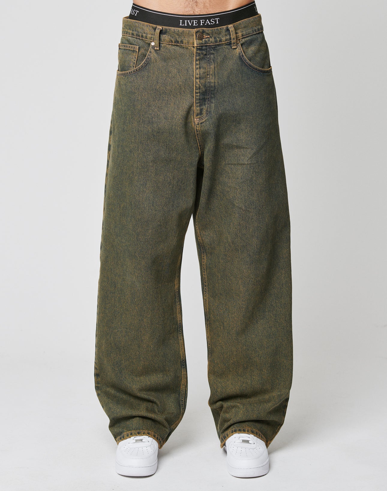 Someone wears LIVE FAST DIE YOUNGās LFDY Baggy Denim in faded greenish-brown with a baggy fit and wide straight leg, paired with white sneakers and black underwear featuring LIVE FAST on the waistband, against a plain white background.