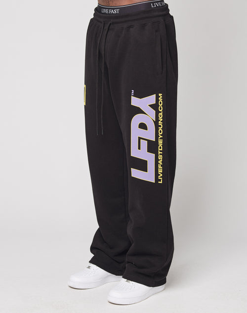 A person wears the LFDY Racing Wide Jogger by LIVE FAST DIE YOUNG, featuring bold white and yellow leg prints, a wide fit, and a broad elastic waistband, paired with white sneakers against a plain light background.