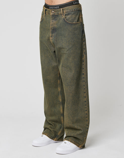 Someone wears LIVE FAST DIE YOUNG’s LFDY Baggy Denim—baggy, faded greenish-brown jeans with wide straight legs and front pockets, made of 100% cotton—showing their black LIVE FAST waistband above the jeans, paired with white sneakers.