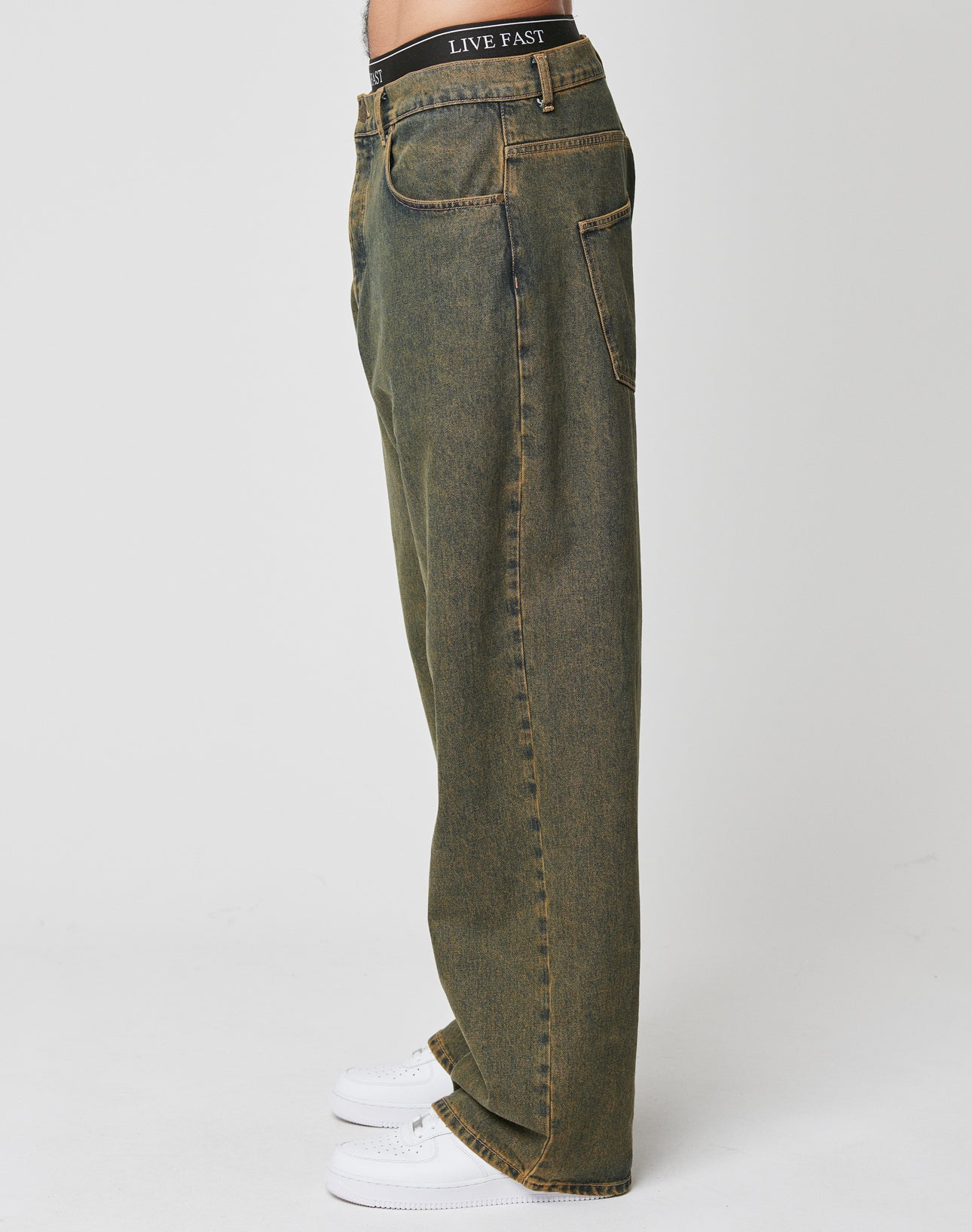 A person stands in profile wearing LFDY Baggy Denim by LIVE FAST DIE YOUNGāloose-fitting, faded olive green jeans with baggy fit, wide straight leg, yellow stitching, back pockets, and a LIVE FAST waistband label. 100% Baumwolle. White background.