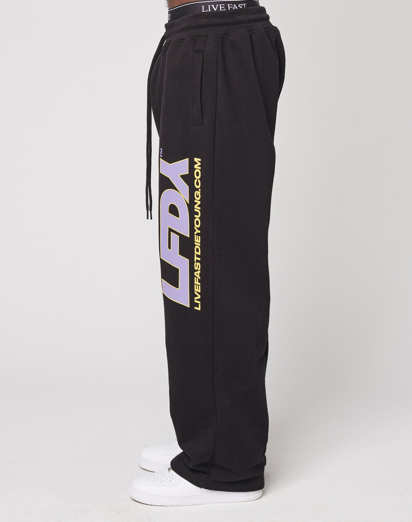 A person wearing black LFDY Racing Wide Jogger by LIVE FAST DIE YOUNG, featuring a wide fit, drawstring waist, Breiter Elastikbund, and bold yellow and white print on the legs, paired with white sneakers.
