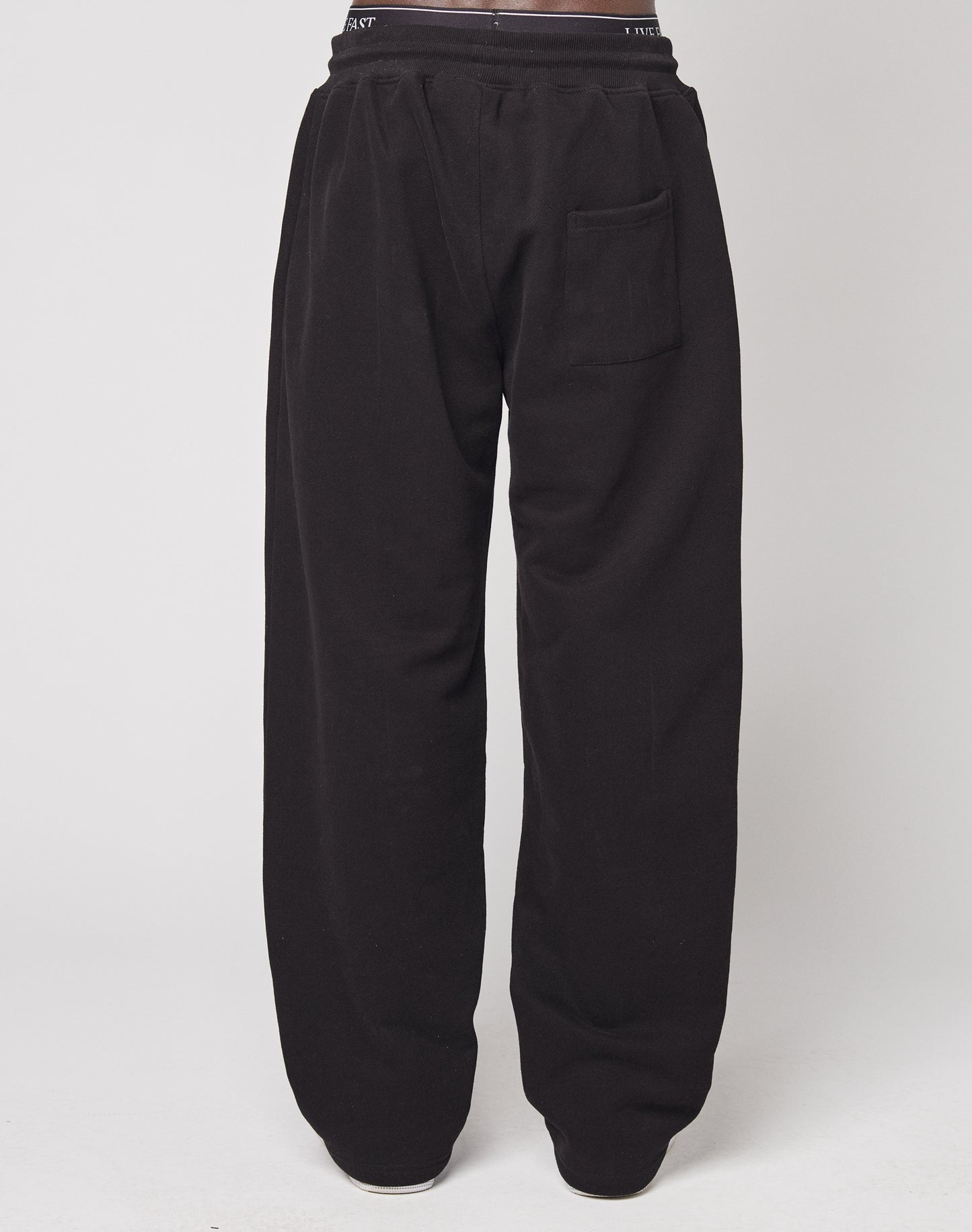 Person trägt LFDY Racing Wide Jogger von LIVE FAST DIE YOUNG in Schwarz mit Elastikbund und einer Gesäßtasche, steht vor weißem Hintergrund und wird von hinten fotografiert.