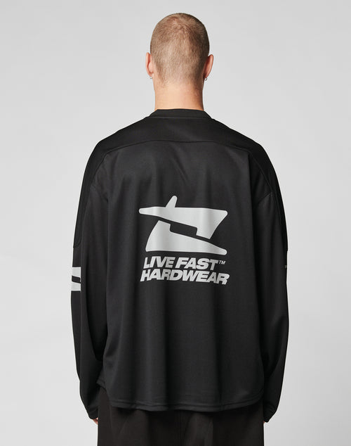 A person with short hair is seen from behind wearing the LIVE FAST DIE YOUNG Live Fast Jersey, an oversized black long-sleeve shirt featuring a bold silver reflective LIVE FAST HARDWEAR print on the back against a plain light gray background.