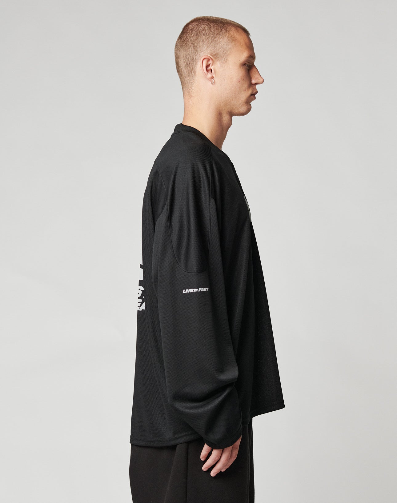 A person with short hair stands in profile against a plain background, wearing the LIVE FAST DIE YOUNG Live Fast Jersey—an oversize black long-sleeve shirt with white reflective print on the sleeve—and dark polyester pants.