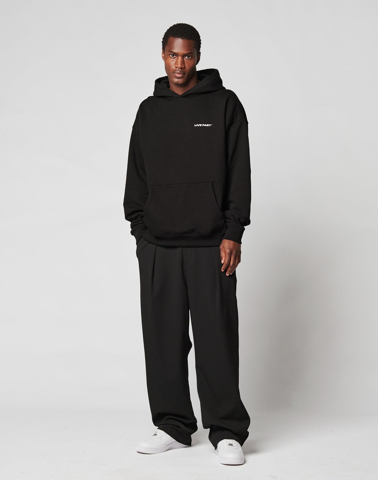 A person stands against a plain light background, wearing a black hoodie with small white text on the left chest, LIVE FAST DIE YOUNG Wide Casual Pants, and white sneakers. They look directly at the camera with a neutral expression.