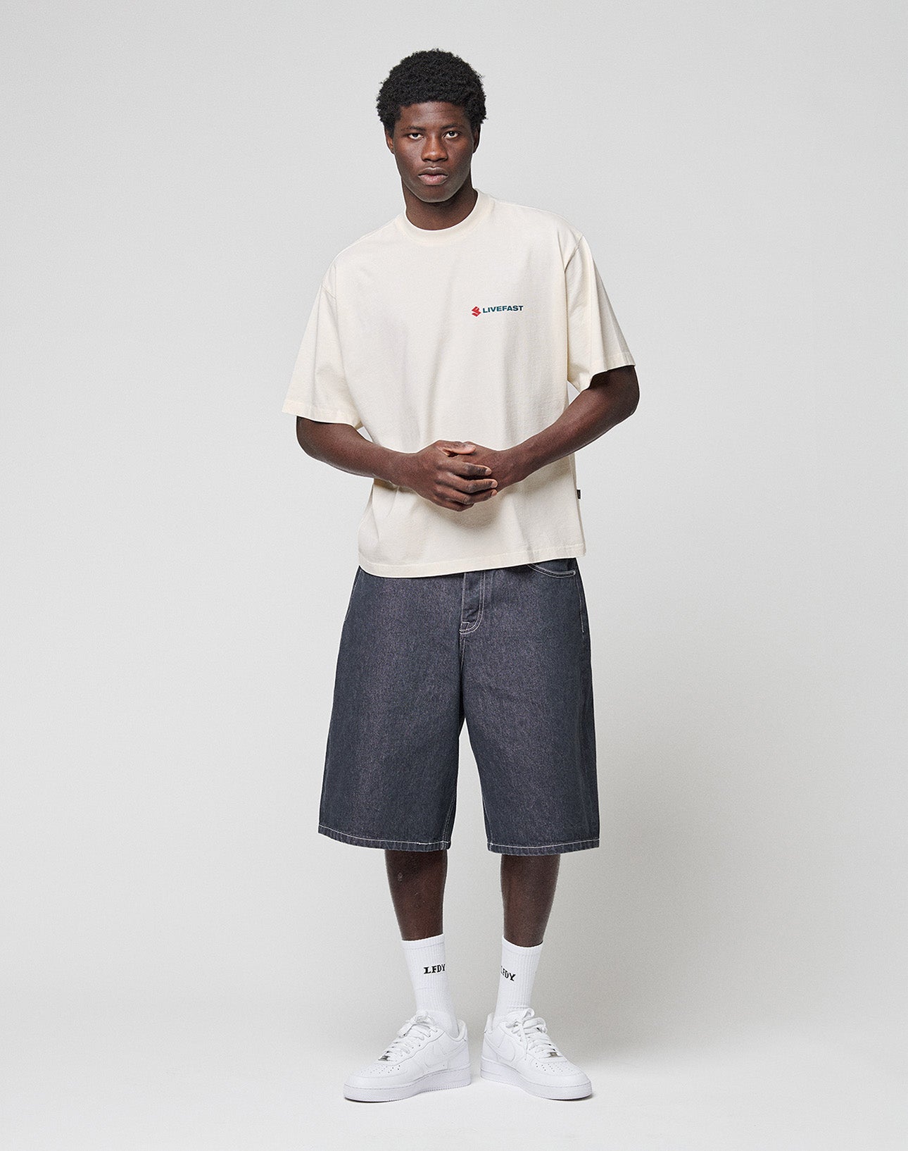 A young man faces forward against a plain background, wearing a baggy cream T-shirt, white crew socks, white sneakers, and dark LIVE FAST DIE YOUNG LFDY Jorts with a Lederpatch. His hands are loosely clasped in front of him.