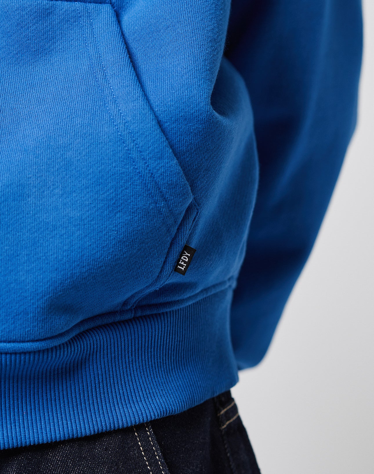 Close-up of a person wearing the LFDY Team Hooded Zip by LIVE FAST DIE YOUNG, a bright blue oversized hoodie with a small black KEN tag near the pocket, paired with dark blue denim jeans.