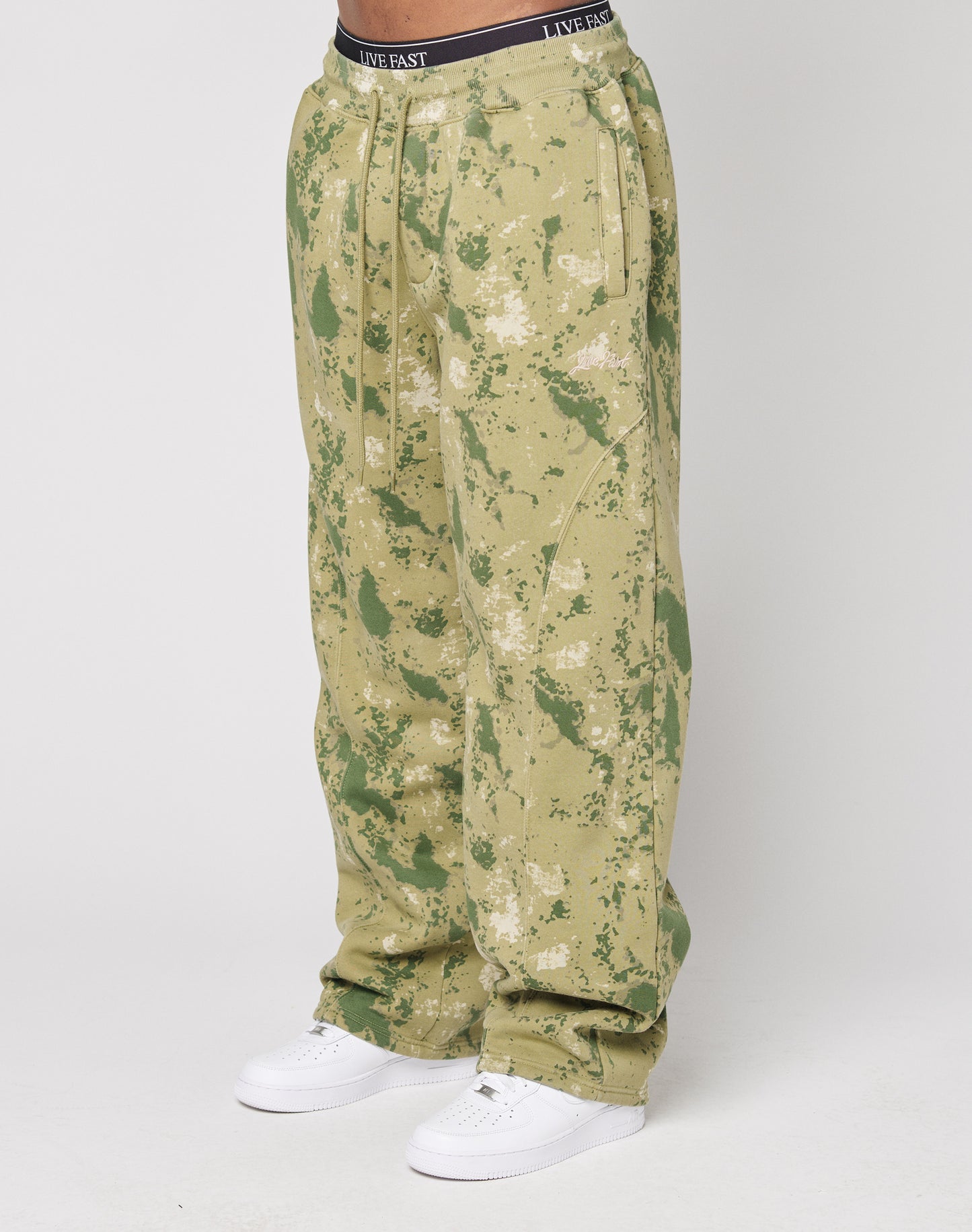 A person wears LIVE FAST DIE YOUNGs Camo Wide Joggerāloose-fitting, green and tan camouflage pants with a wide fitāpaired with white sneakers and a visible black LIVE FAST waistband, set against a plain white background.