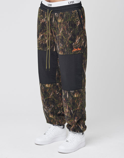 A person wears LFDY Fleece Pants by LIVE FAST DIE YOUNG, featuring camo print, black knee patches, nylon panels, Leisure Paris orange embroidery, drawstring waist, elastic cuffs, and an oversized fit—paired with white sneakers.