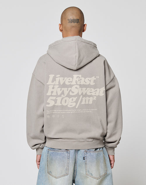 A person with a shaved head, wearing the LIVE FAST DIE YOUNG Basic 510 Hooded Zip in Oversize Fit with bold white “Live Fast Hvy Sweat 510g/m²” text on the back, stands facing away from the camera, paired with light blue jeans.