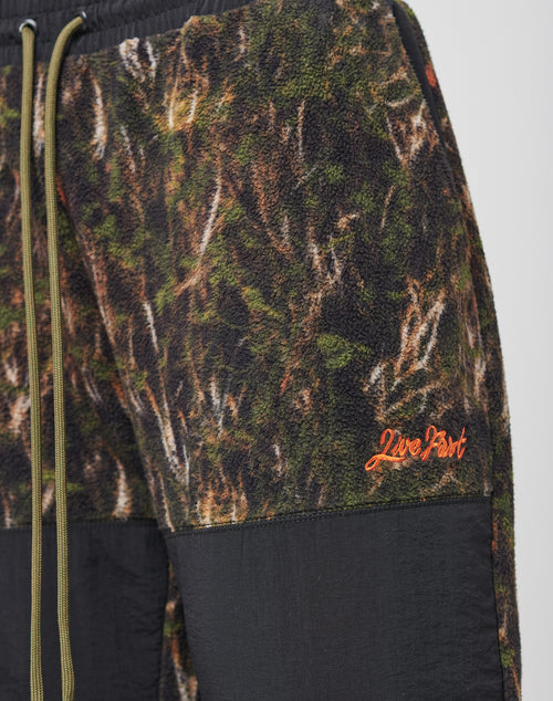 Close-up of LFDY Fleece Pants by LIVE FAST DIE YOUNG, featuring a green/brown camo fleece, black nylon panels, orange Live Fast embroidery near the pocket, olive green drawstrings, and an oversized fit for comfort.
