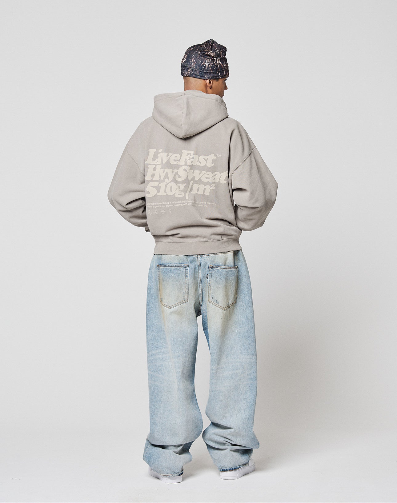 A person faces away, wearing the LIVE FAST DIE YOUNG Basic 510 Hooded Zip—an oversize gray hoodie with front and back prints—paired with light blue baggy jeans, a dark patterned headscarf, and white shoes against a plain background.