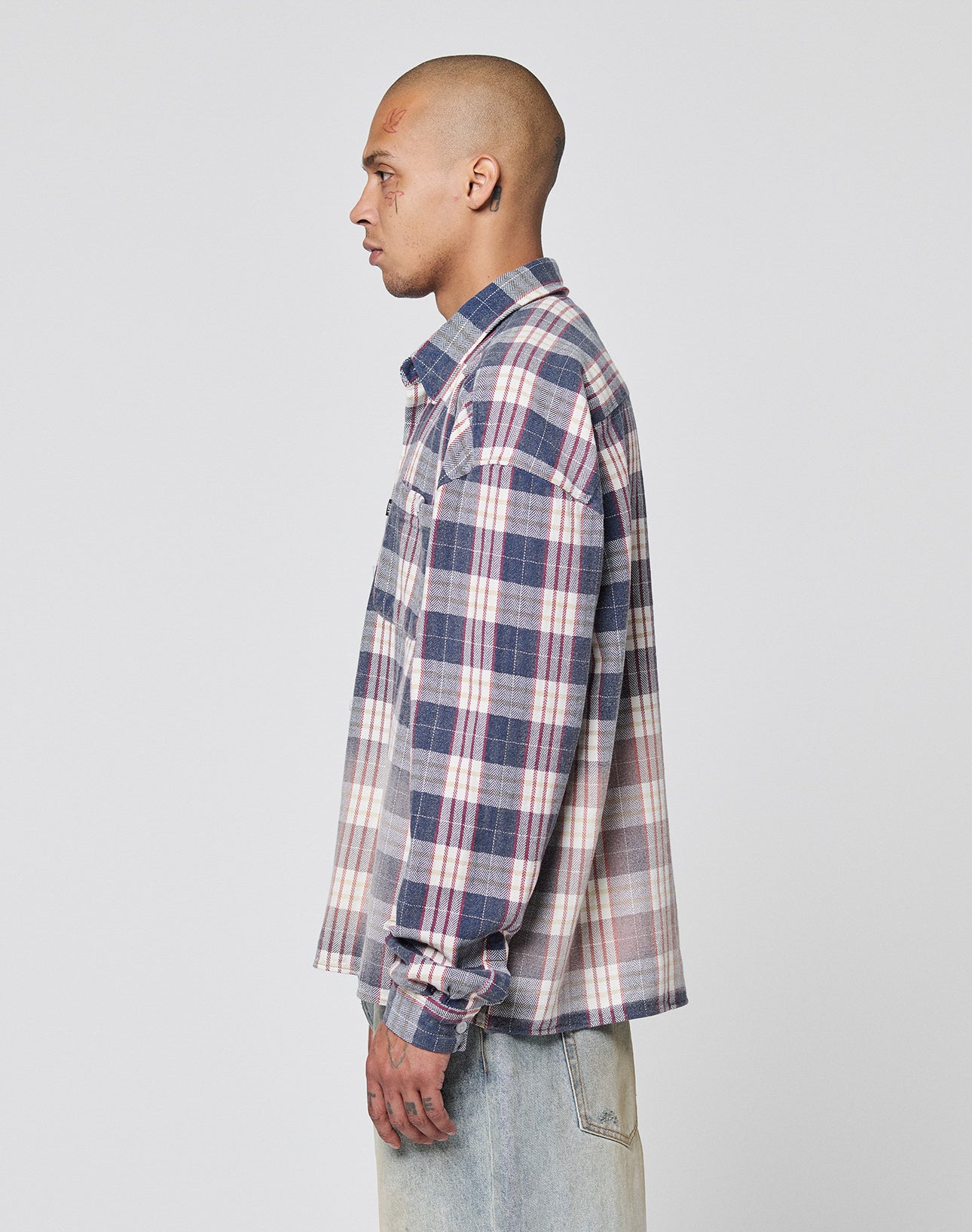 A person with a shaved head wears the LIVE FAST DIE YOUNG Vintage Flannel in blue, white, and pink plaid and light-wash Oversize Fit jeans, posing in profile against a plain white background.