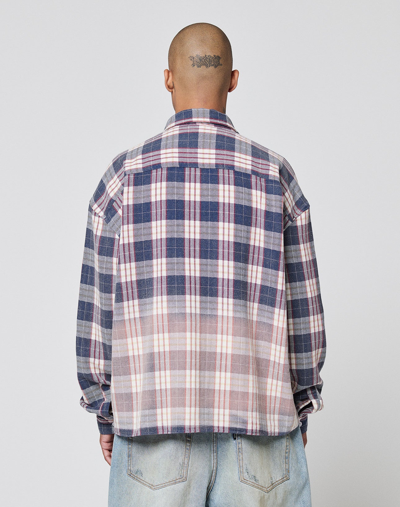 A person with a shaved head and tattoo is seen from behind wearing the LIVE FAST DIE YOUNG Vintage Flannel, an oversized blue, white, and pink plaid shirt with zweiteiliger Hemdkragen, paired with light blue jeans.