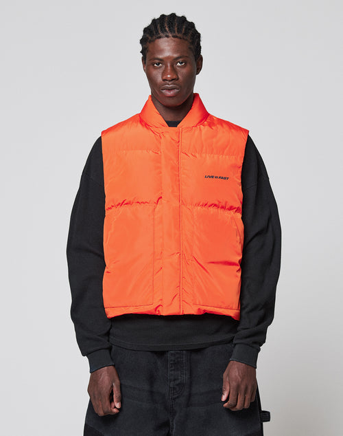 A man wears the LIVE FAST DIE YOUNG Live Fast Cropped Vest in orange with Druckknöpfe over a black sweatshirt and dark pants, standing against a plain light gray background and looking directly at the camera.