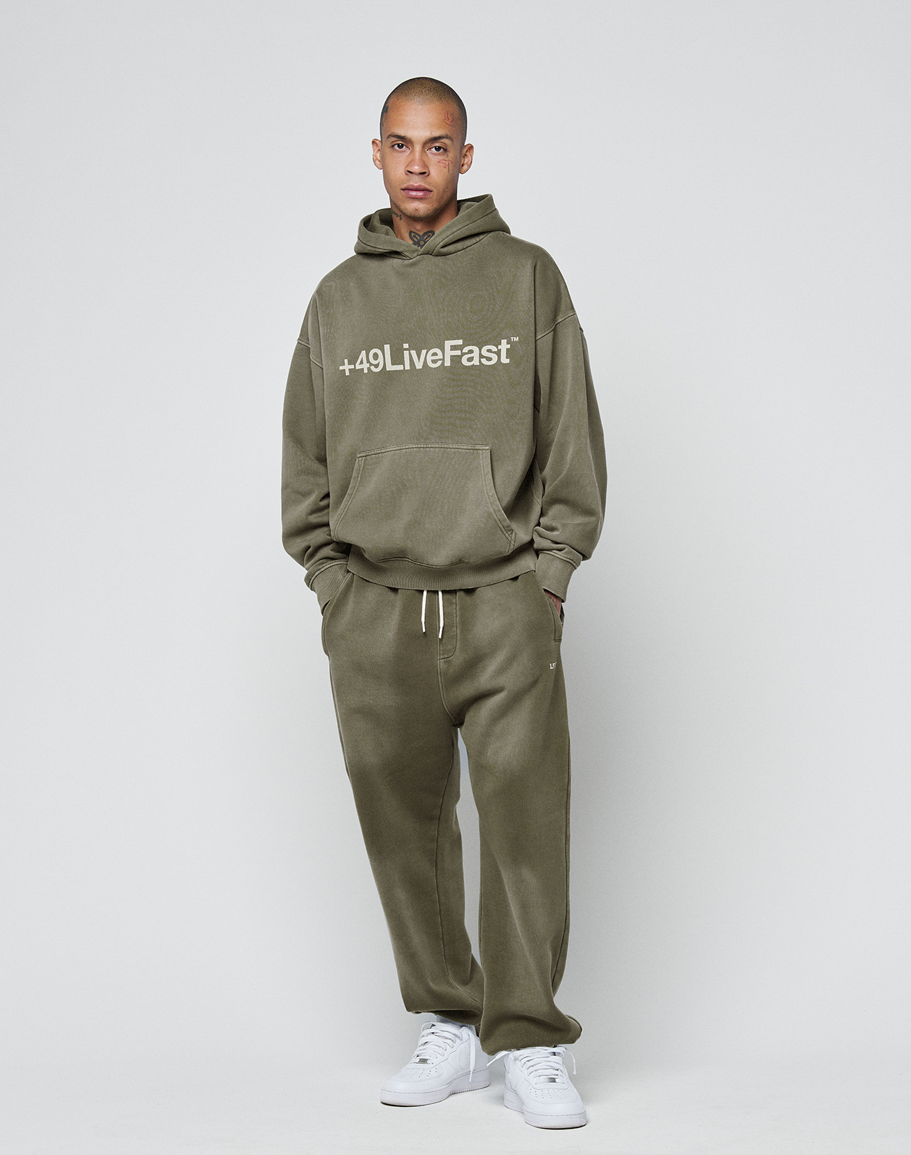 A person stands against a plain background wearing the LIVE FAST DIE YOUNG Basic +49 Hooded in olive green with a handmade fade effect, paired with matching sweatpants and white sneakers for an oversize fit, hands in pockets and a neutral expression.