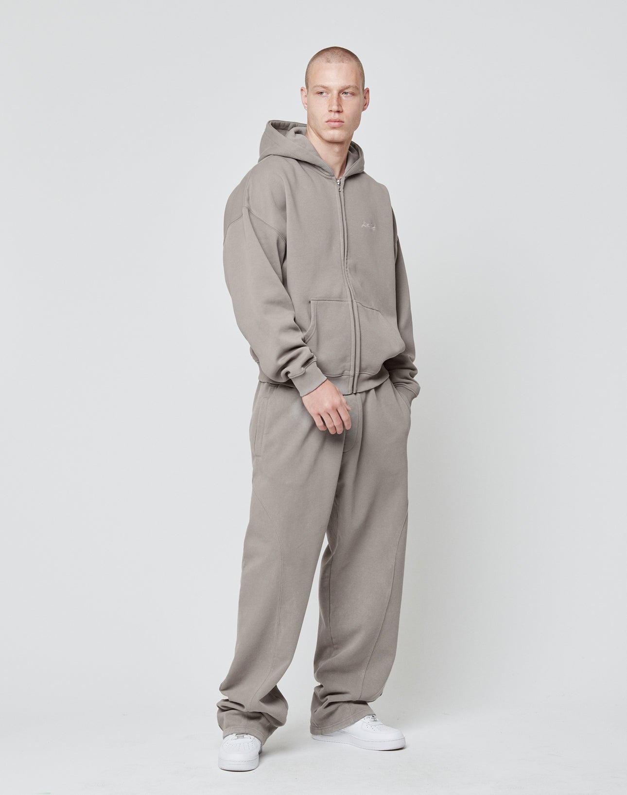 A man stands against a plain white background wearing LIVE FAST DIE YOUNGs Basic Wide Jogger and matching taupe hoodie, both made from soft cotton with an elastic waistband. His hands are in his pockets as he looks slightly to the side.