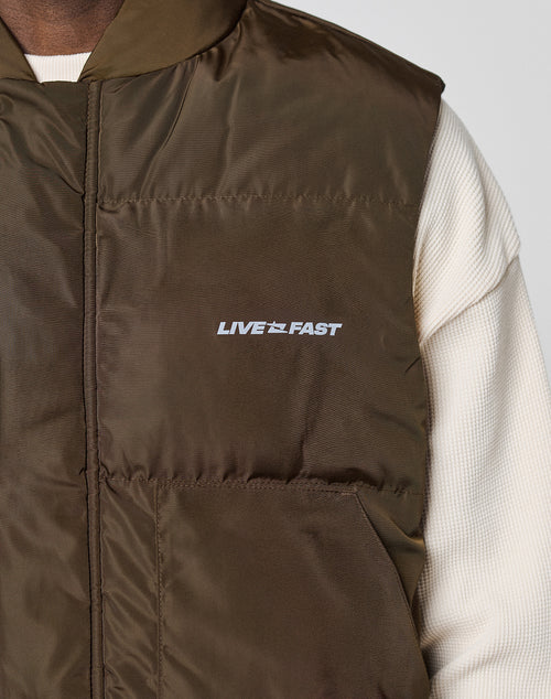 A close-up shows a person wearing the Live Fast Cropped Vest by LIVE FAST DIE YOUNG—an olive green, polyester puffer vest with “LIVE FAST” printed in white on the chest, layered over a cream long-sleeve shirt.