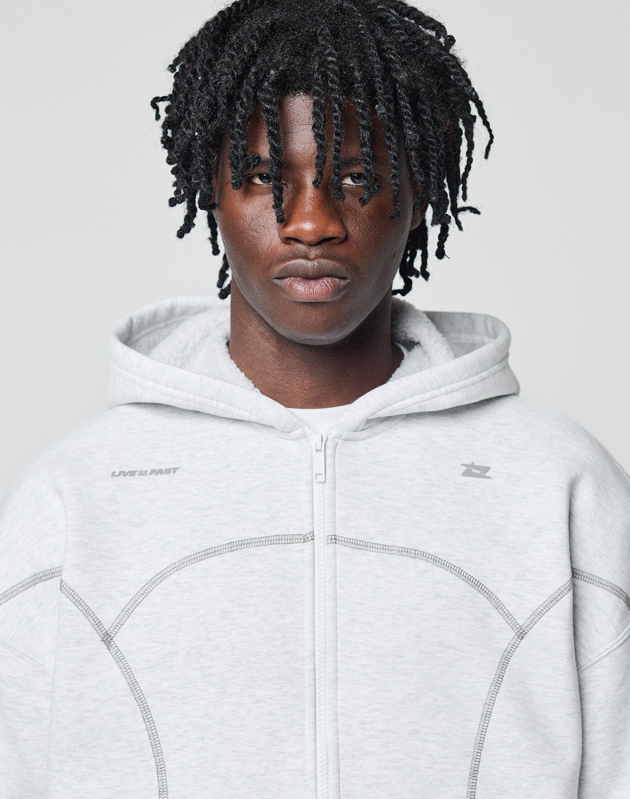 A young person with twisted black hair wears the LFDY Active Fleece Hooded Zip by LIVE FAST DIE YOUNG—a light gray, oversized zip-up hoodie with thin curved seams and a minimal logo on the shoulders, set against a plain light background.